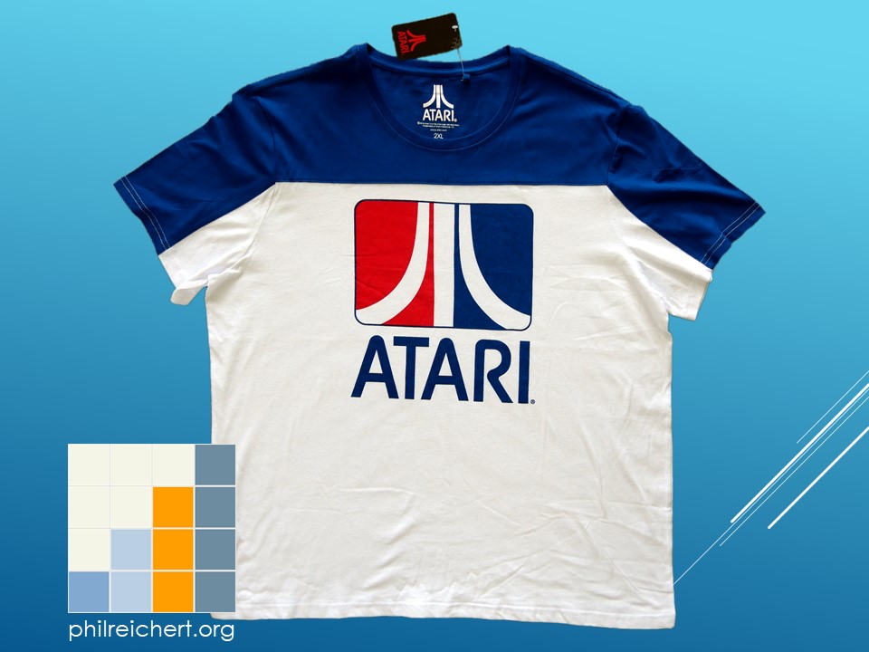 Front view of an Atari-branded retro T-shirt laid flat, showing the bold central Atari logo and contrasting blue, white, and red colour scheme